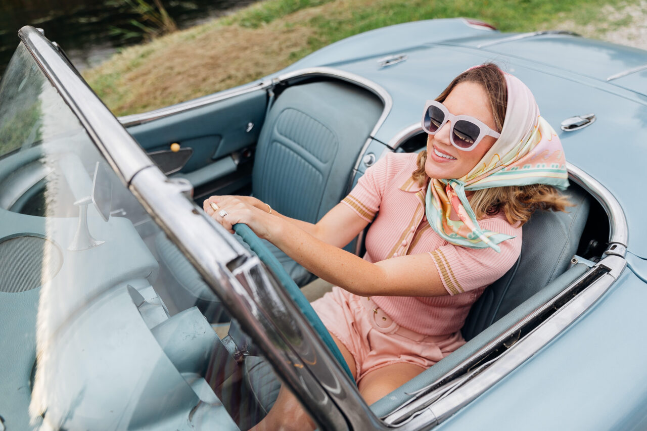 Woman in sunglasses drives a vintage car, wearing a colorful scarf and pastel outfit, inspired by The Scout Guide.